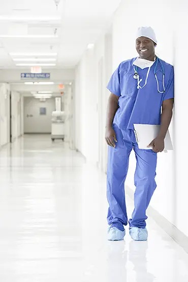 A smiling healthcare professional in blue scrubs leans against a wall in a hospital corridor, holding a clipboard.