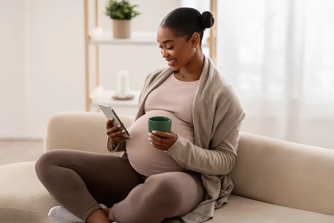 A pregnant woman sits on a couch, smiling while holding a phone and a green cup, dressed in comfortable clothing.