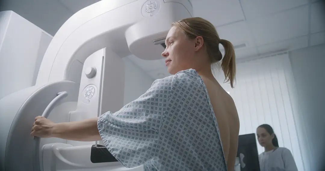 A woman in a hospital gown is preparing for a medical procedure near a machine, with a healthcare worker in the background.