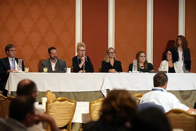 A panel of speakers at a conference, seated behind a table, discussing in front of an audience.