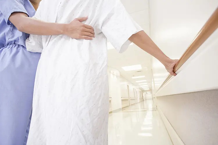 A caregiver supports a patient in a hospital corridor, both wearing medical attire. The setting is bright and clinical.