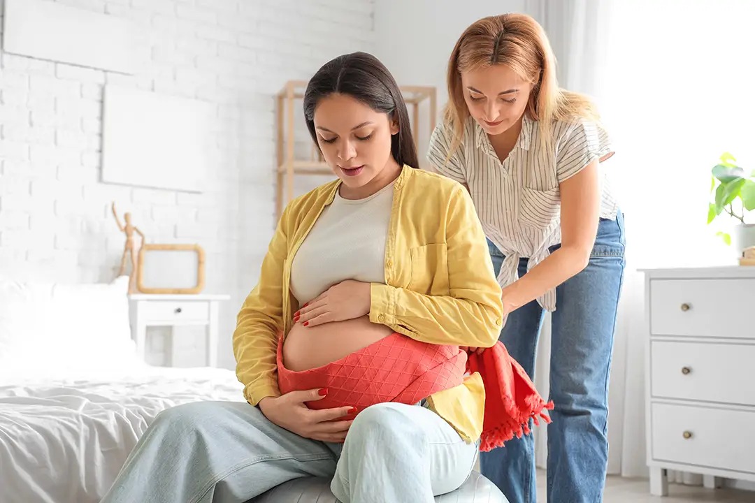 A pregnant woman sits on a ball, gently holding her belly, while another woman adjusts a support wrap around her waist.