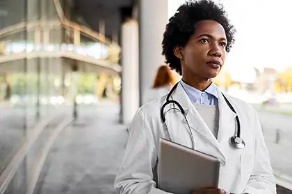 A thoughtful doctor in a white coat holds a tablet, standing outside a modern building with reflective glass.