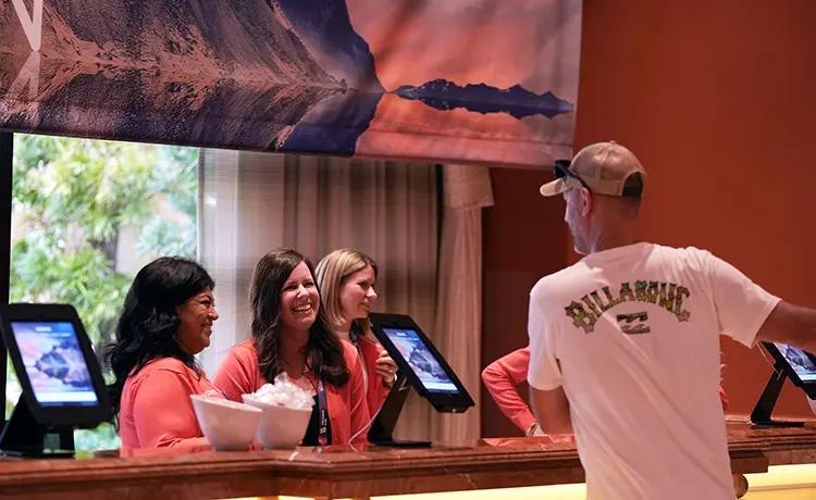 A hotel reception scene with three smiling staff members at a counter, engaging with a guest. Tablets are visible on the counter.