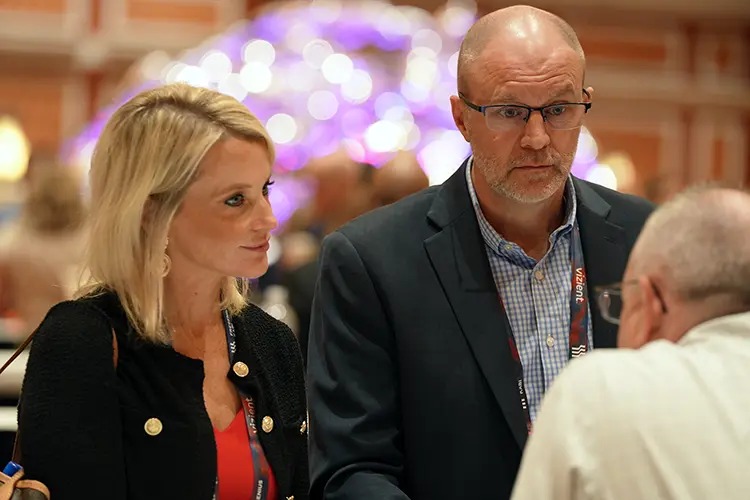 A man and a woman engage in conversation at an event, with a blurred background of lights and attendees.