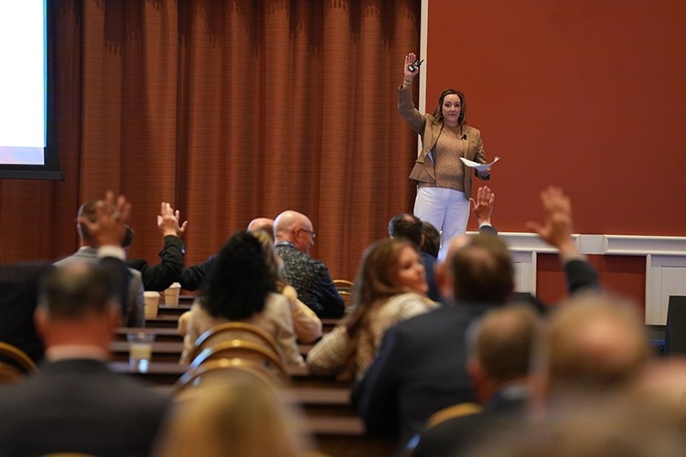 A speaker stands on stage with one hand raised, engaging an audience that is responding with raised hands in a conference setting.