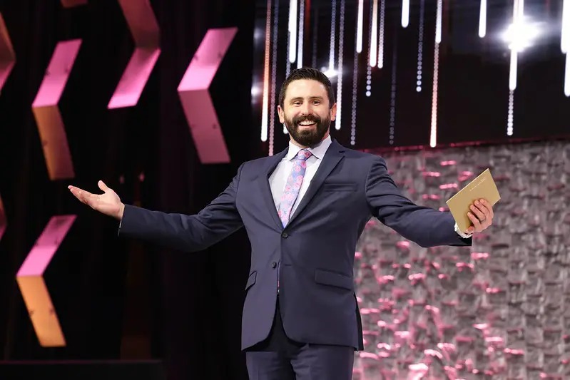 A man in a suit stands on stage with arms outstretched, smiling, holding a notebook, against a colorful backdrop.