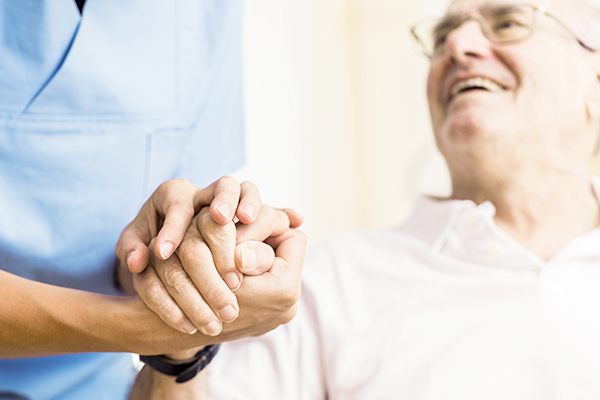 A caregiver holds hands with an elderly man, both smiling, conveying warmth and connection.