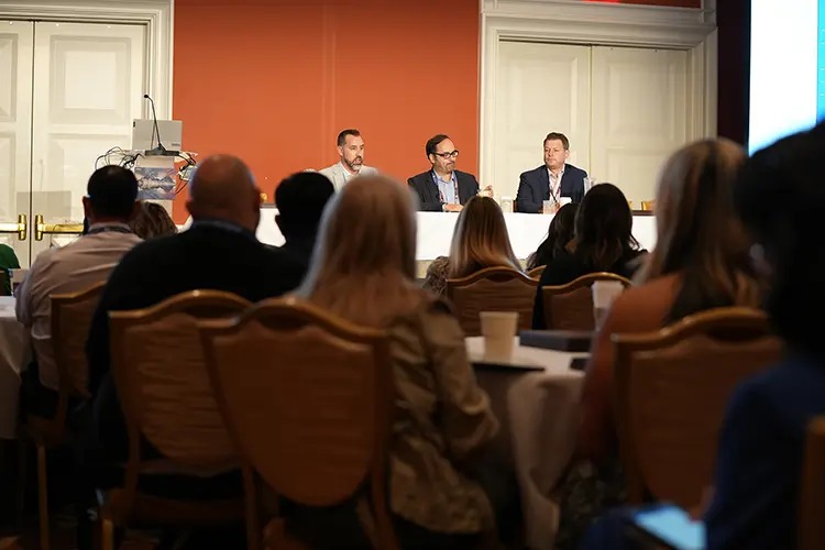 A panel discussion featuring three speakers at a conference, with an audience visible in the foreground.