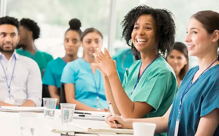 A group of healthcare professionals in scrubs engaged in a meeting, with one woman smiling and speaking.