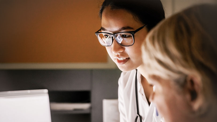 A female healthcare worker with a stethoscope and glasses looking at a laptop screen with a colleague.